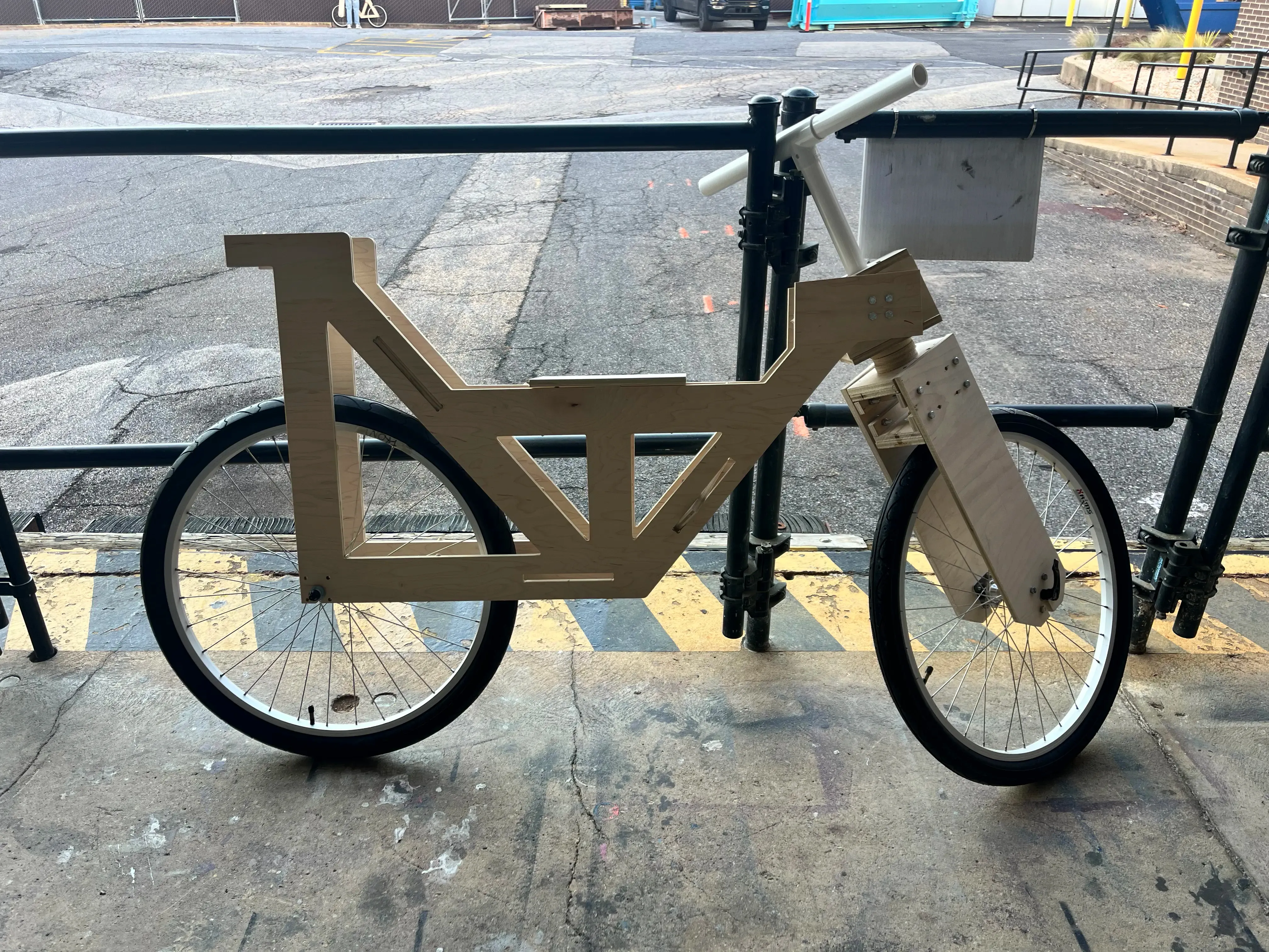 Assembled wooden-frame bicycle prototype locked to a bike rack outside a lab building